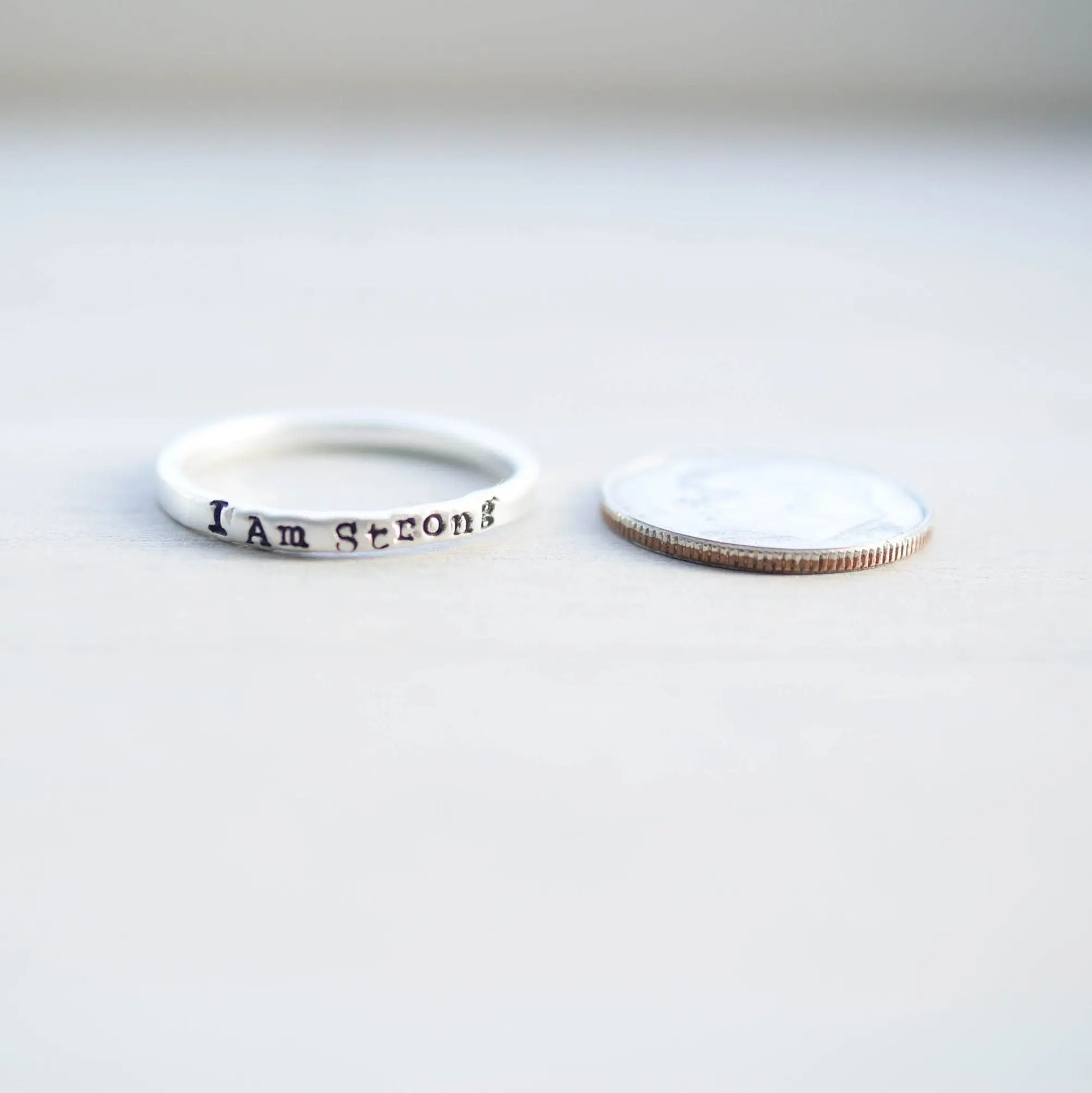Sterling Silver stacking ring handstmaped with I Am Strong next to dime